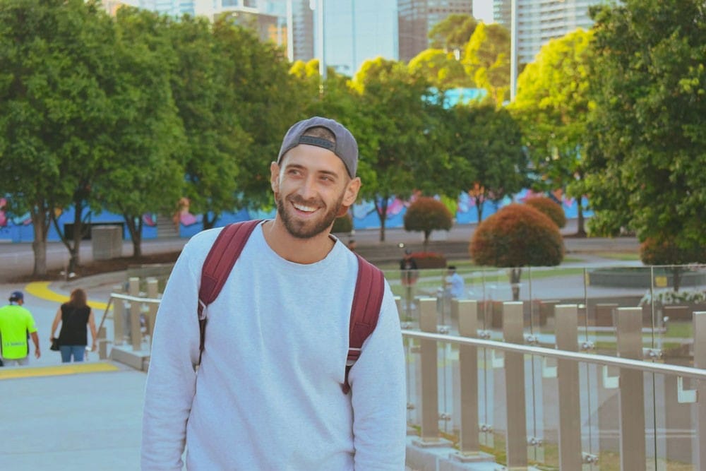 Vivir en Brisbane chico con una gorra sonriendo a cámara y detrás un parque y la ciudad